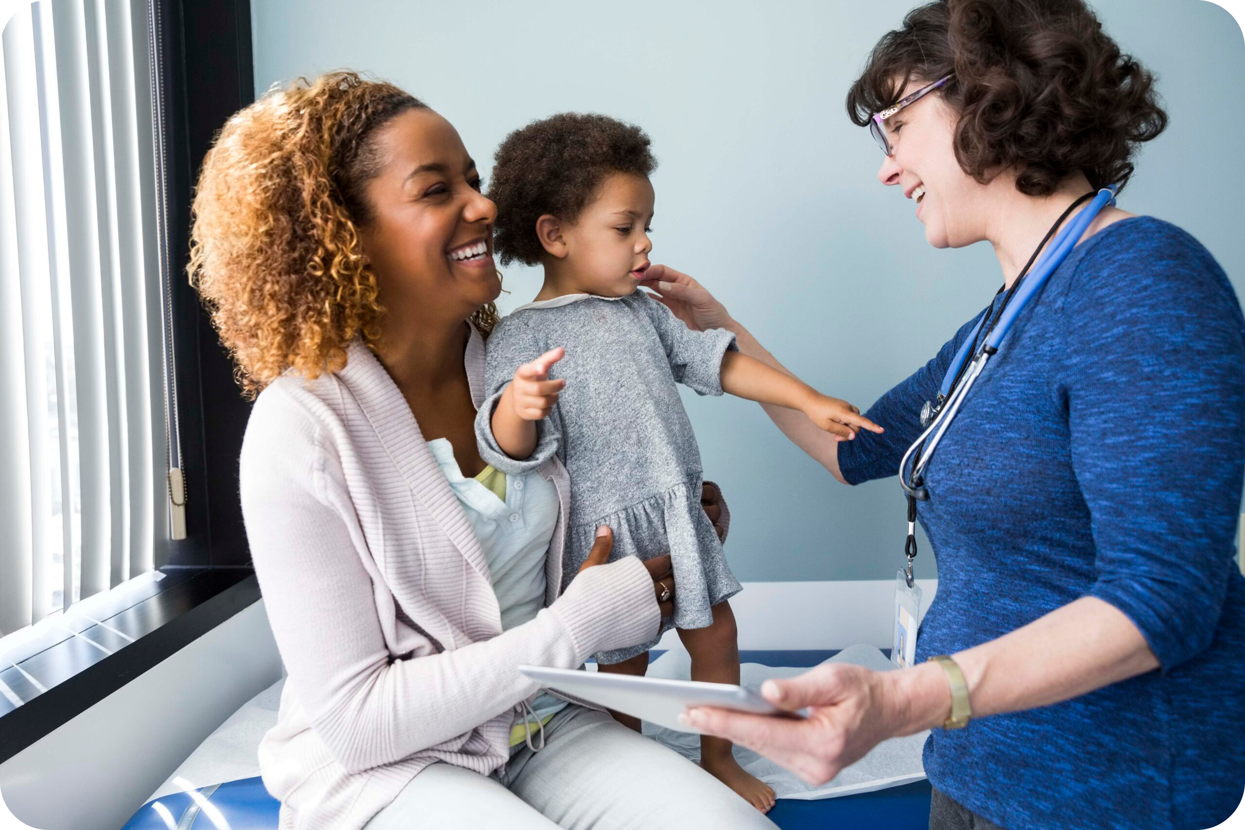 Smiling White female pediatrician greets a toddler and her Black mother in an exam room.